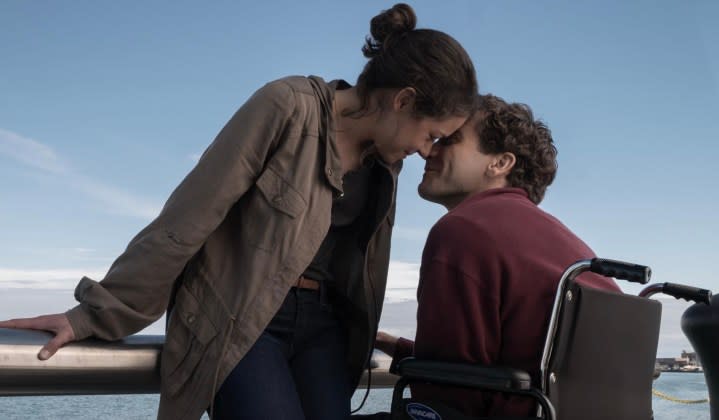 A woman puts her head against a man's head who is sitting in a wheelchair.