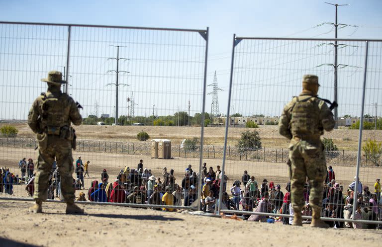Migrantes esperan junto a una valla fronteriza mientras son vigilados por miembros de la Guardia Nacional de Texas, para ingresar a El Paso, Texas, el miércoles 10 de mayo de 2023. (AP Foto/Andres Leighton)
