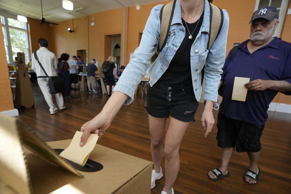 A person in denim jacket over dark shirt and shorts places a piece of paper into a ballot box