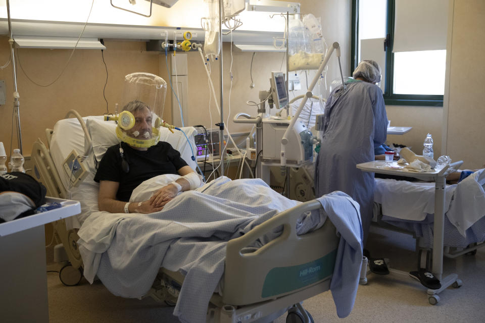 60-year-old Patient Nazzareno Santilli, breathes under oxygen CPAP (continuous positive air pressure) headgear ventilation looks on as Dr. Elisabetta Teti encourages his bed neighbor in a sub-intensive COVID-19 unit of the Tor Vergata Polyclinic Hospital, in Rome, Saturday, Nov. 7, 2020. Communication is challenging when patients wear oxygen helmets and Teti is covered by layers of protective gear. (AP Photo/Alessandra Tarantino)