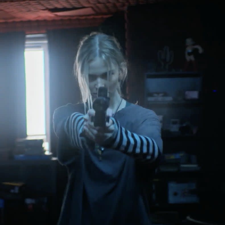Sasha Luss aiming a gun with two hands in a dimly lit room with blinds on the windows
