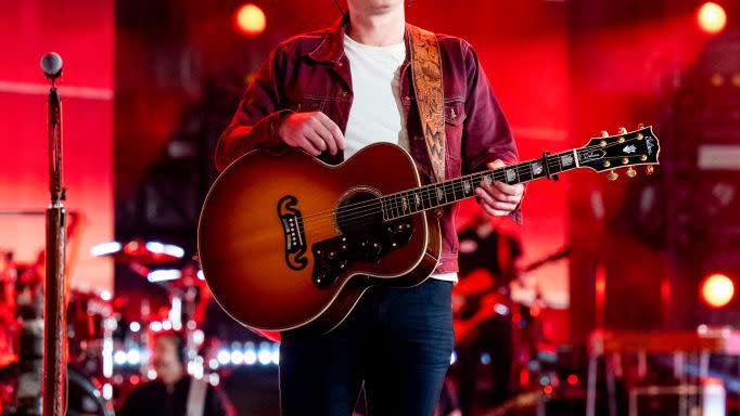 morgan wallen stands on a stage with a guitar strapped around him, he looks up to the right and wears a backward ball cap, maroon collared shirt, white t shirt and jeans