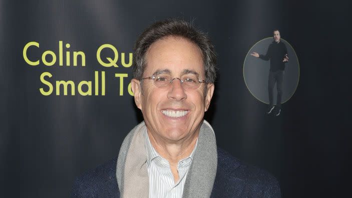 jerry seinfeld smiling for a photo at a premiere event wearing a suit jacket and scarf