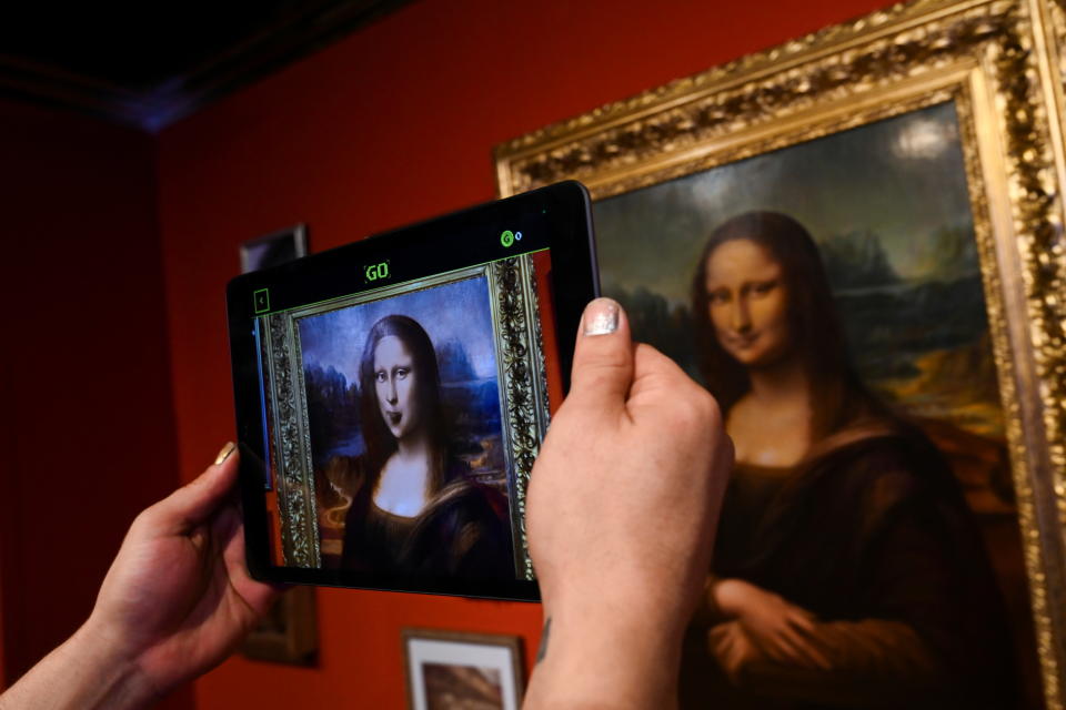 A tablet displays an animated NFT artwork of the Mona Lisa with a replica of the original in the background.