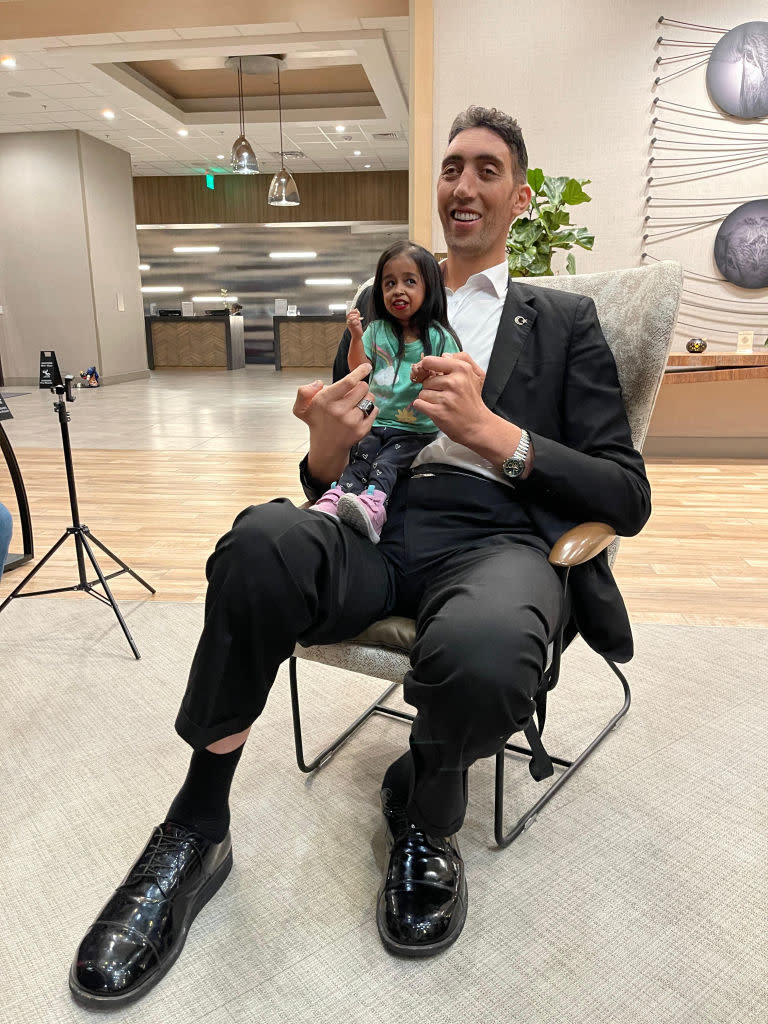 Man in a suit holding a small, lifelike doll, both seated and gesturing 'peace' signs