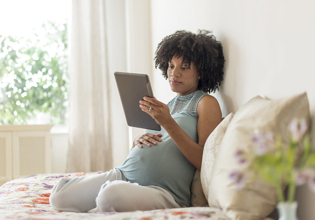 A pregnant Black woman sits on her bed by a window, looking at her tablet.