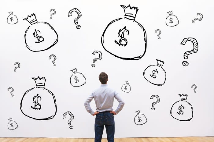 Man with hands on hips looking at a wall covered with drawings of money bags and question marks