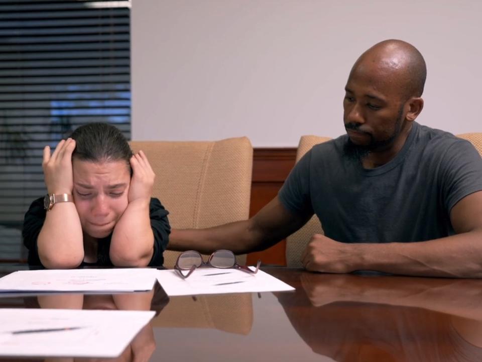 natalia grace sits at a table with her head in her hands looking at several papers. antwon mans sits next to her with a hand on her back