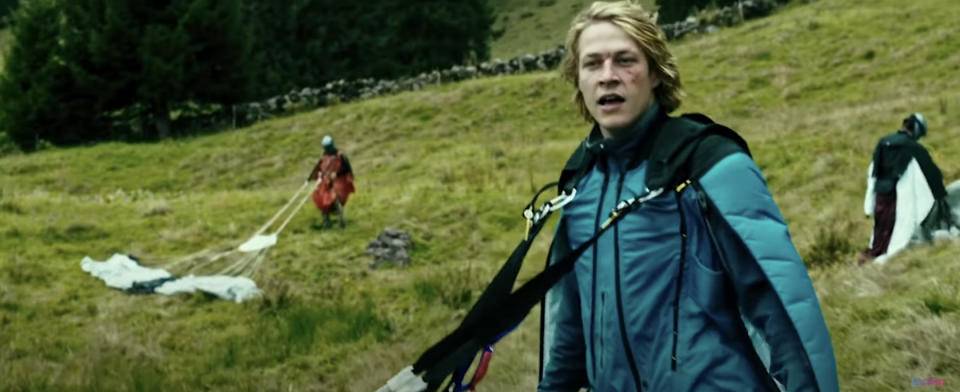 Man in a blue jacket looks surprised in a grassy field with others and parachutes in the background