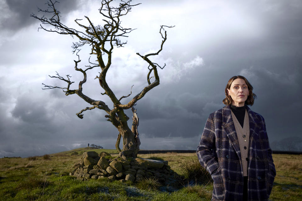 An image for Suranne Jones: Investigating Witch Trials showing Suranne Jones standing in a field. There is a small tree behind her, its branches twisted and gnarled, and there are no leaves on the tree. The sky behind her is full of dark clouds