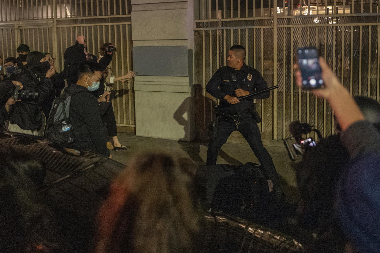 A police officer draws back his baton inside an open gate surrounded by a group of demonstrators, some of whom are using their cellphones to shoot video of him.