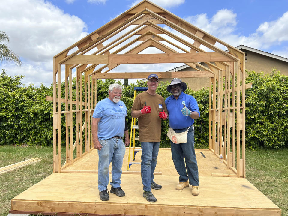 In this photo provided by Amikas, from left, Amikas Vice President Herb Kelly, El Cajon City Councilmember Steve Goble, and Pastor Rolland Slade celebrate on the front porch of cabin four, almost halfway framed behind them at Meridian Baptist Church in El Cajon, Calif., on April 16, 2022 Each cabin has 96 square feet of enclosed, secured living space and 48 square feet of covered porch to foster a sense of community. (Lisa Kogan/Amikas via AP)