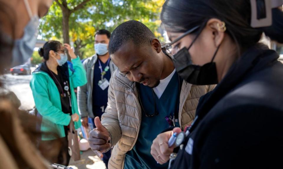 A Black man with trim hair and moustache leans over a clipboard held by a younger Latina woman wearing glasses and a face mask, with green trees and other people around them.