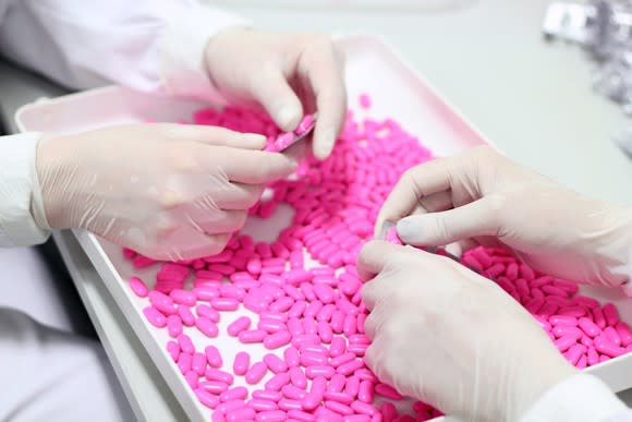 Two sets of gloved hands press a pile of pink pills into packages.