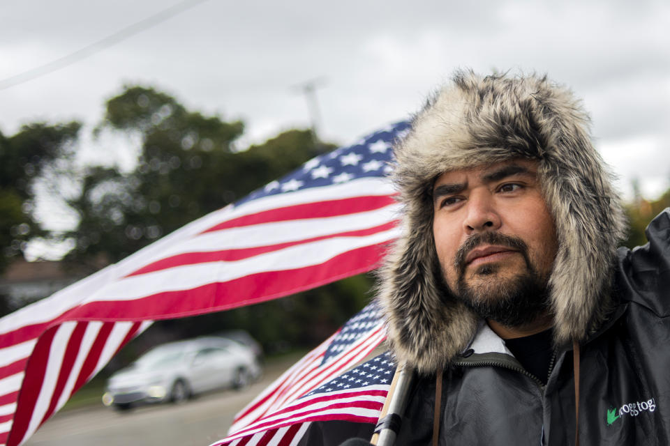 Michael Perez, 48 of Saginaw, stand with waving American flags to show solidarity alongside other General Motors' Flint Assembly Plant employees and UAW members, who line the street with picket signs on the 31st day of the nationwide UAW strike against GM after stalled contract negotiations on Wednesday, Oct. 16, 2019, outside of the Flint Assembly Plant in Flint, Mich. Perez has been employed as an electrician in the body shop for more than 10 years. ÒWe get to keep our job and our health insurance, but we're mainly fighting for the younger crowd and the future of this plant," Perez said. (Jake May/The Flint Journal via AP)