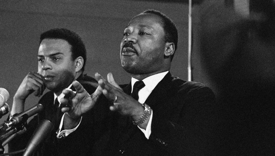 In this Feb. 7, 1968, file photo, Andrew Young, left, stands beside Dr. Martin Luther King Jr. as he speaks to an audience and promises a massive demonstration in the spring in Washington, D.C.