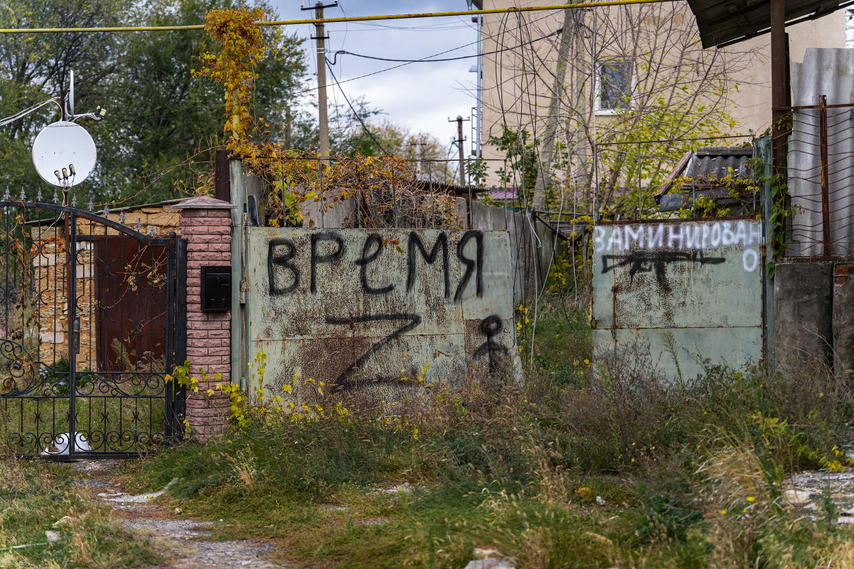 An overgrown walkway leads to a gate flanked by a concrete wall with spray-painted inscriptions in Russian.
