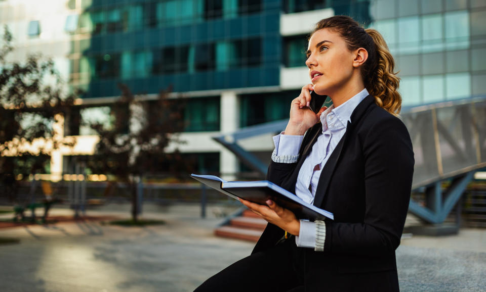 Successful businesswoman or entrepreneur taking notes and talking on cellphone outdoors. City business woman working.