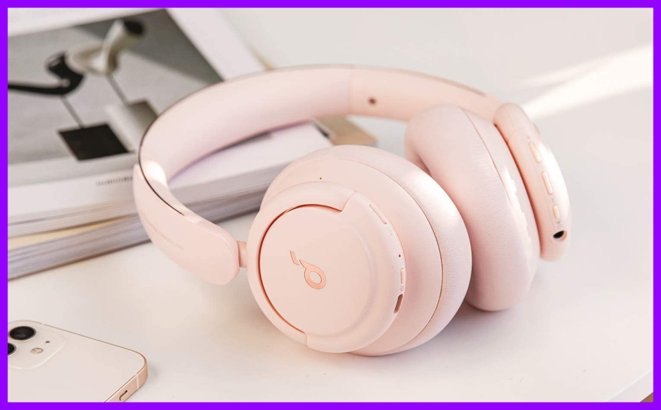 Pink wireless headphones laying atop a desk near a phone and magazine. 