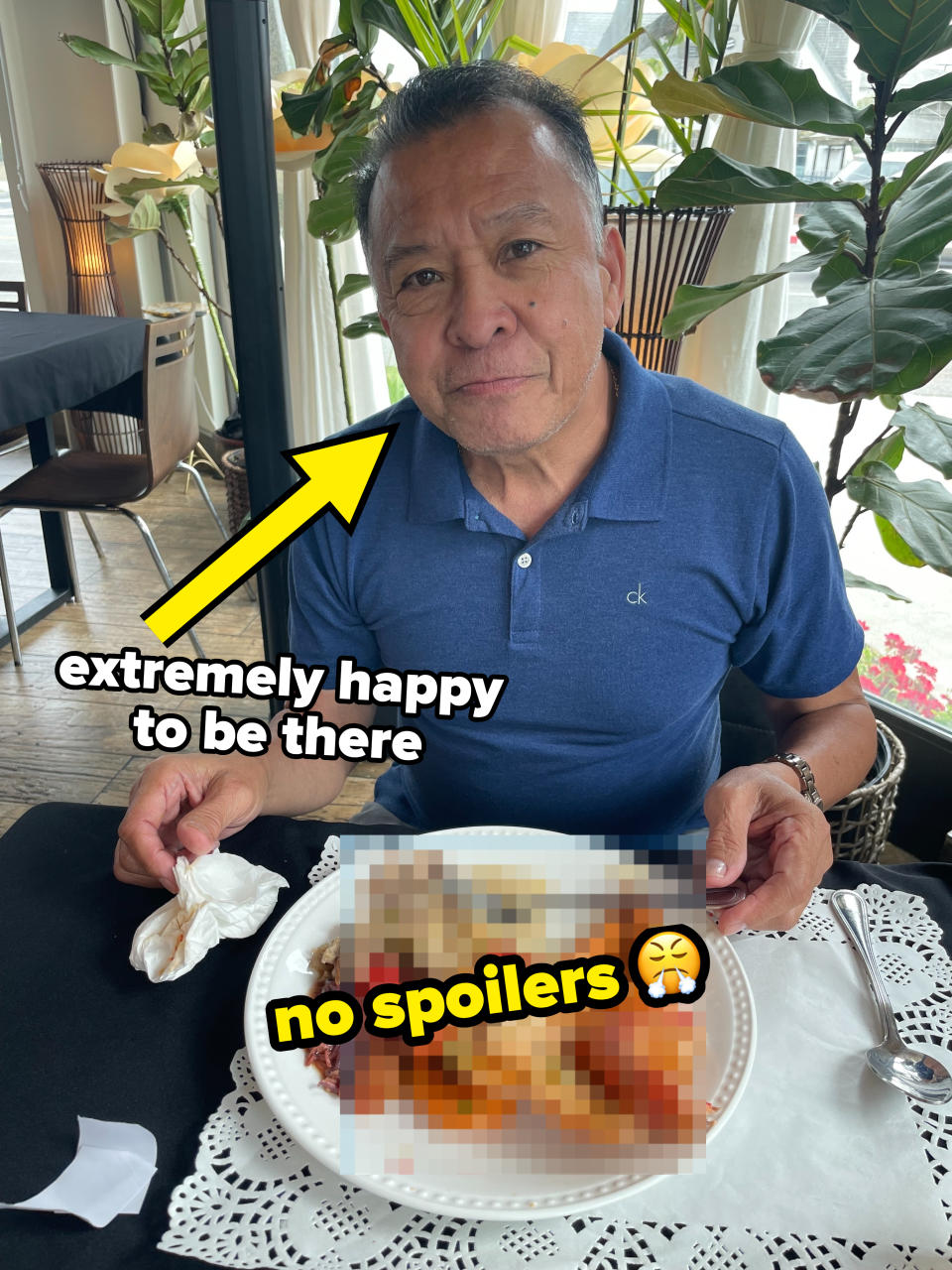 A man in a blue polo shirt at a restaurant holding a fork and napkin, seated in front of a white plate filled with various foods