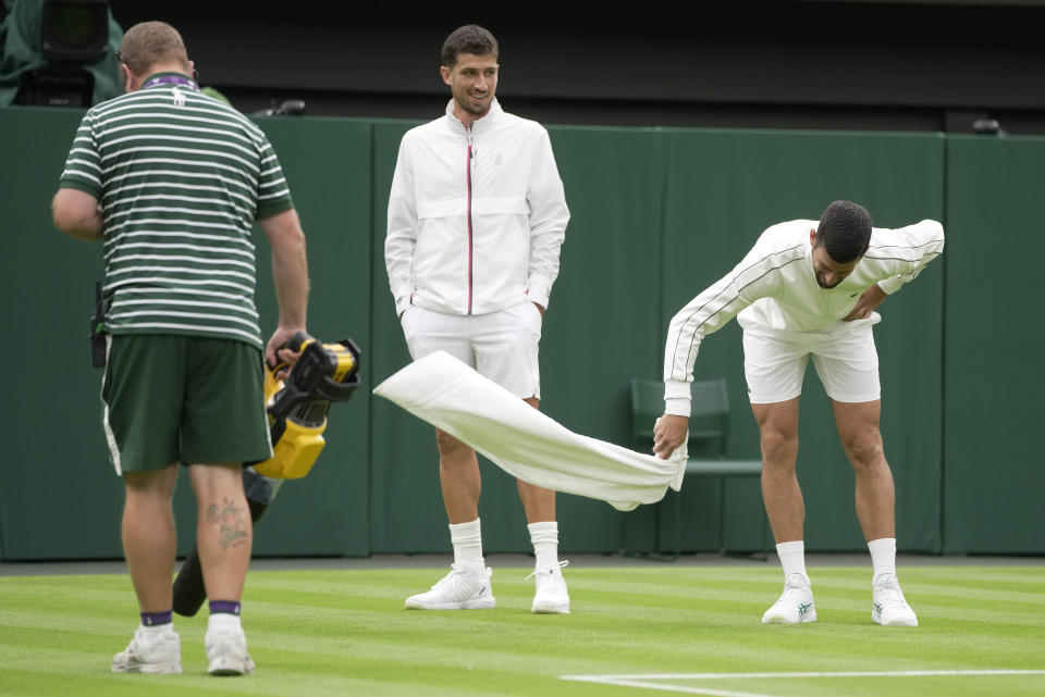 A member of the ground staff uses a leaf blower and Serbia's Novak Djokovic wafts his towel to assist with drying the court as Argentina's Pedro Cachin looks on after a rain break in their first round men's singles match on day one of the Wimbledon tennis championships in London, Monday, July 3, 2023. (AP Photo/Kin Cheung)