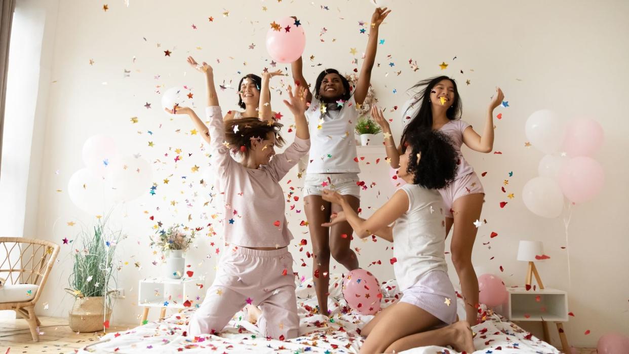 multiethnic women wearing pyjamas dancing on bed under falling confetti