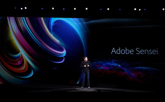 A man on a darkened stage with a colorful background with the words Adobe's Sensei, the company's AI system.