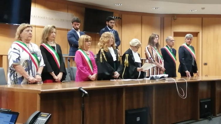 Los jueces con cintas oficiales se paran detrás de un escritorio de madera en una sala del tribunal con paneles.