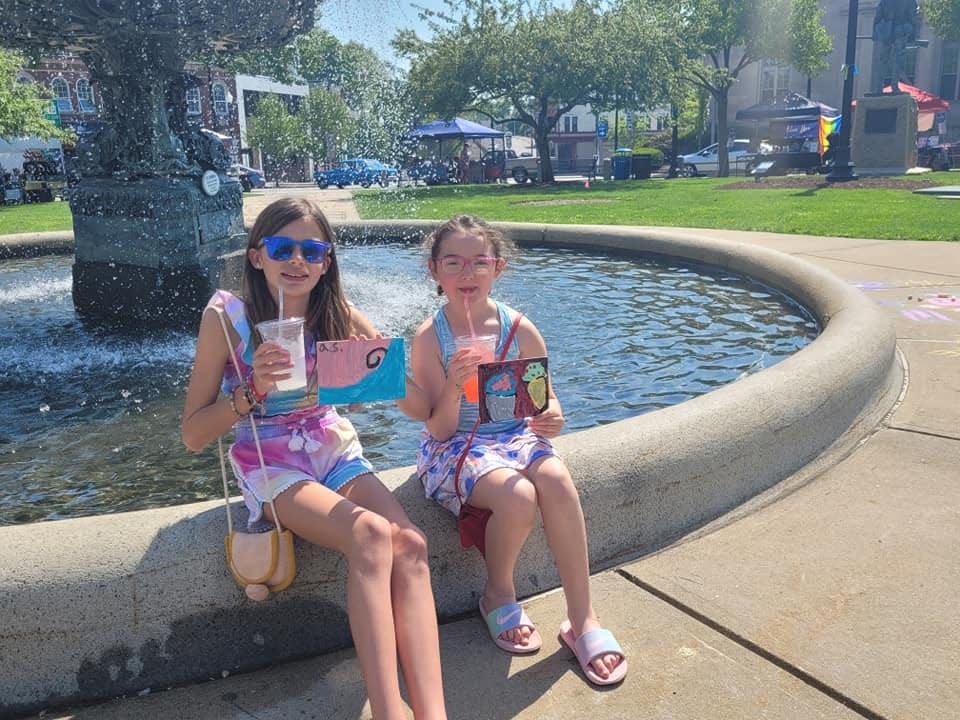 Aubrie Shaw, 10, left, and Shealyn Hughes, 8, right, sip some cool drinks and show off their artwork at the second annual Downtown Art Jam on Taunton Green on Saturday, May 21, 2022.