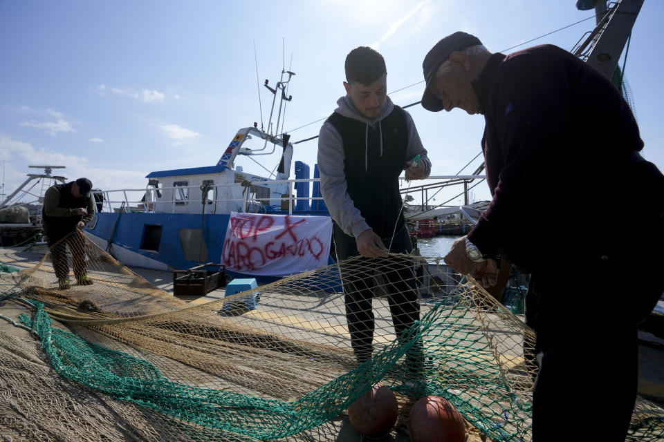 Fishermen mend nets in front of his fishing boat with a banner reading "stop for gasoline increase", in the Roman port of Fiumicino, Friday, March 11, 2022. Fishermen, facing huge spikes in oil prices, stayed in port, mending nets instead of casting them. Nowhere more than in Italy, the European Union’s third-largest economy, is dependence on Russian energy taking a higher toll on industry.(AP Photo/Andrew Medichini)