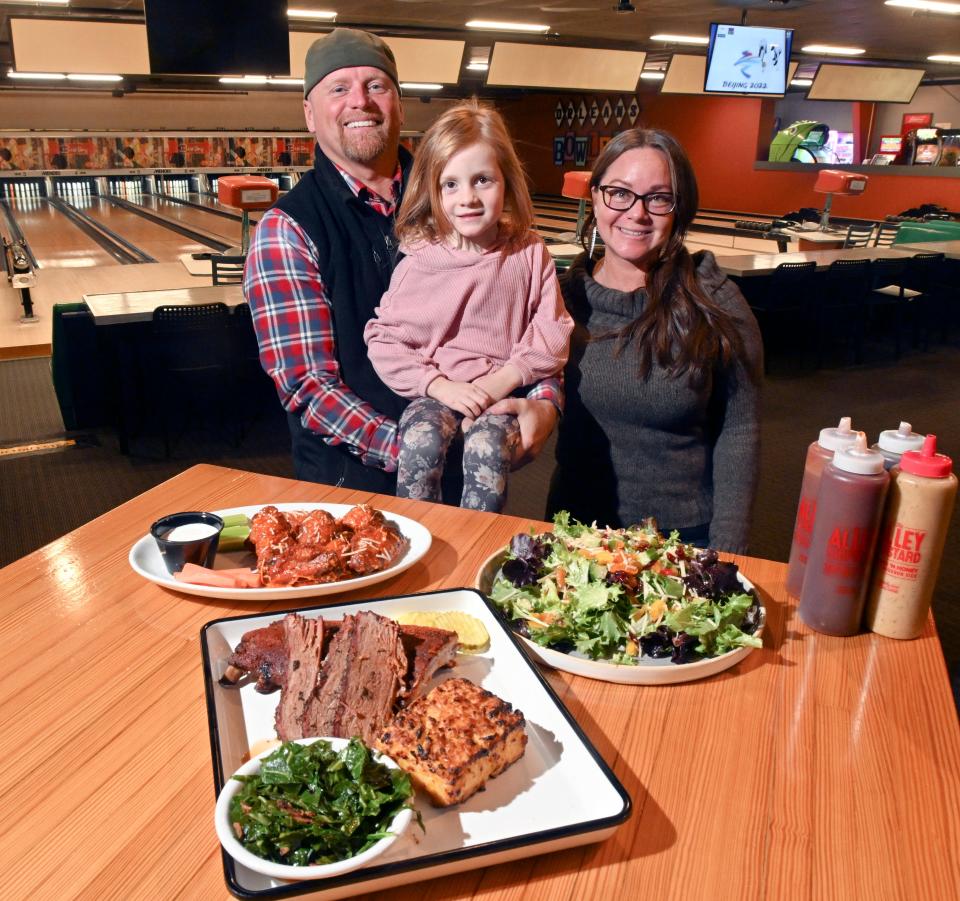 Marion Rebecca Currier is joined by her parents and owners of The Alley Bowling & BBQ Brook Carlson and Dave Currier with some of the restaurant's food; Brisket, ribs and corn pudding , farm salad, wings and collard greens in this 2022 photo.