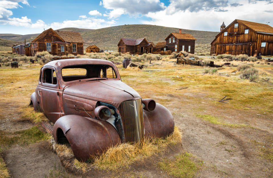 Rusty, old car sits in an abandoned, grassy ghost town with dilapidated wooden buildings in the background. The setting appears desolate and historical