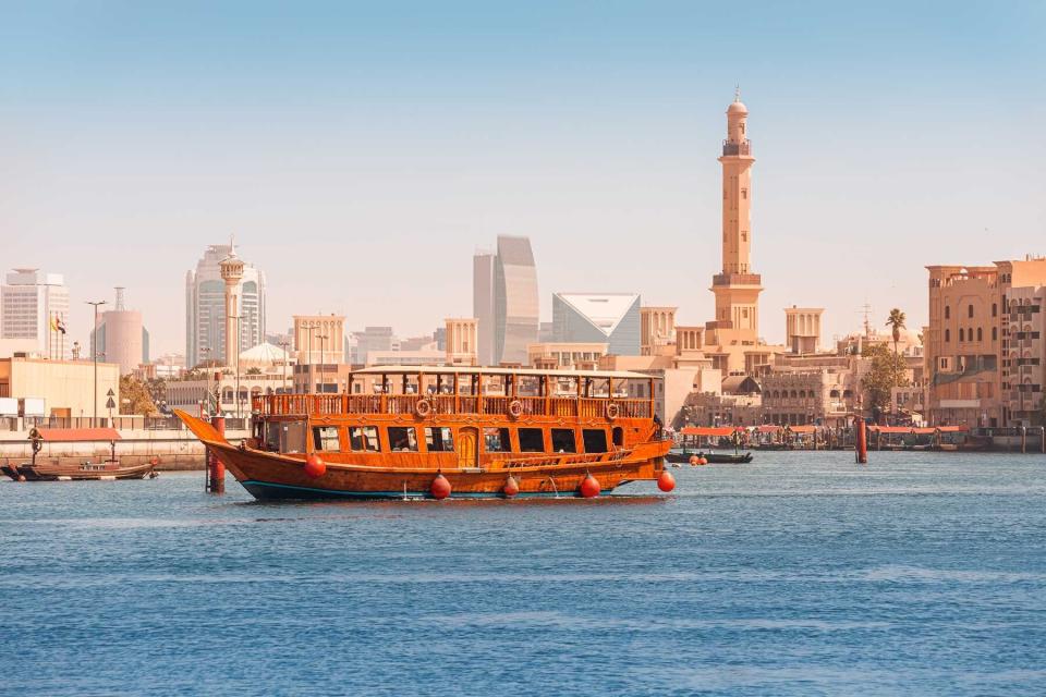 An authentic wooden cruise ship cruises and transports tourists along the Dubai Creek Canal with skyscrapers and minarets of mosques in the background. Travel and sightseeing