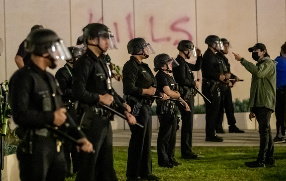 Los Angeles police officers stand shoulder to shoulder wearing riot gear. A protester stands in front of them pointing at them.