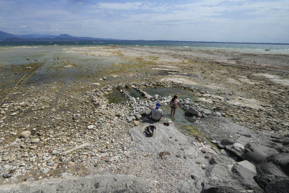 A child with a man stand on the peninsula of Sirmione, on Garda lake, Italy, Friday, Aug. 12, 2022. Lake Garda water level has dropped critically following severe drought resulting in rocks to emerge around the Sirmione Peninsula. (AP Photo/Antonio Calanni)