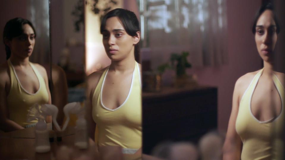 Woman in a yellow halter top reflected in a mirror, looking thoughtful. The setting appears to be a bedroom with soft lighting. Names unknown