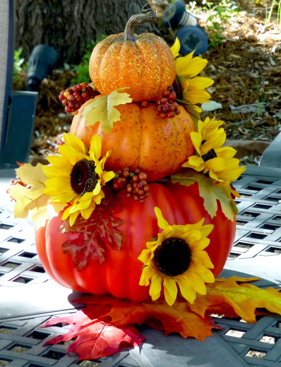 A showy pumpkin stack of three pumpkins of graduated sizes ornamented with seasonal sunflowers, berries, and fall colored leaves make an eye-catching centerpiece.