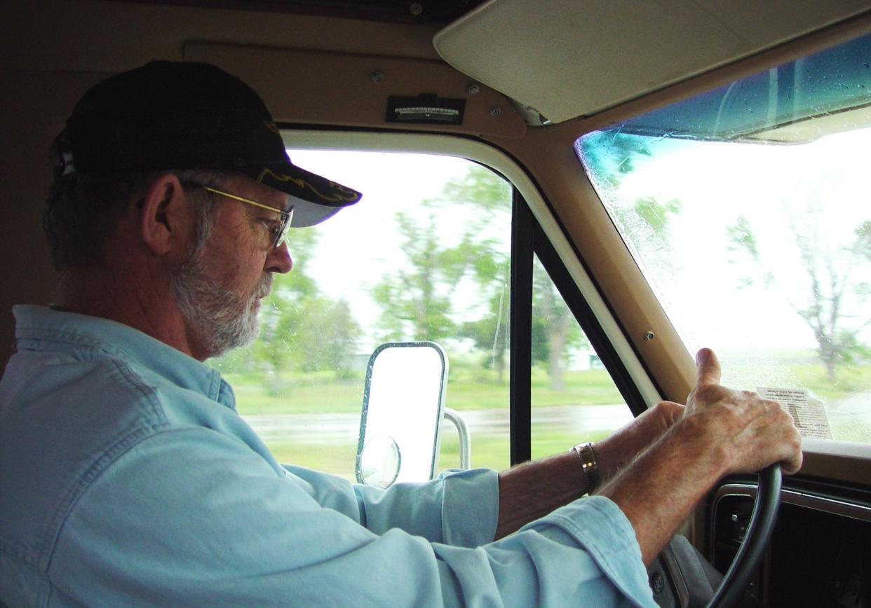 A man driving a RV across Texas in the rain. He is wearing a ball cap and blue denim shirt.