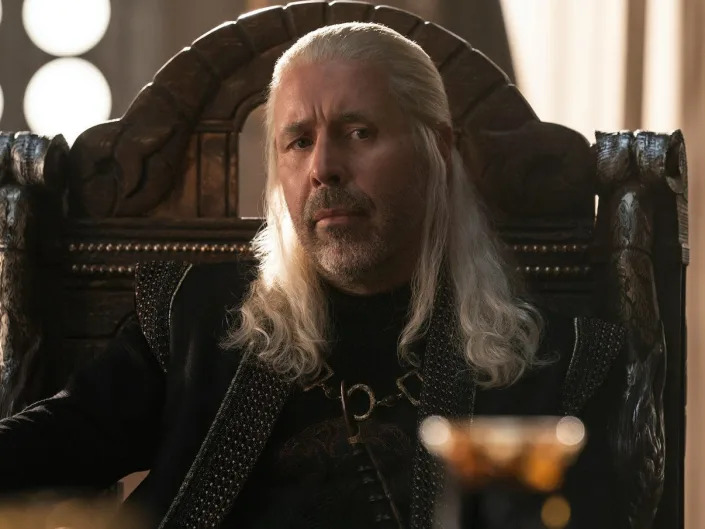 A man with long silvery hair sitting in a regal chair, looking contemplative.