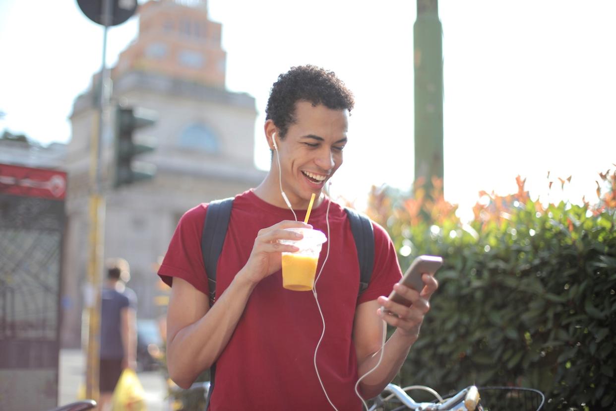100 Perfect pick up lines for guys that want to make their date laugh and smile. Pictured: A man wearing earbuds with a beverage in one hand smiling at his phone in the other hand.
