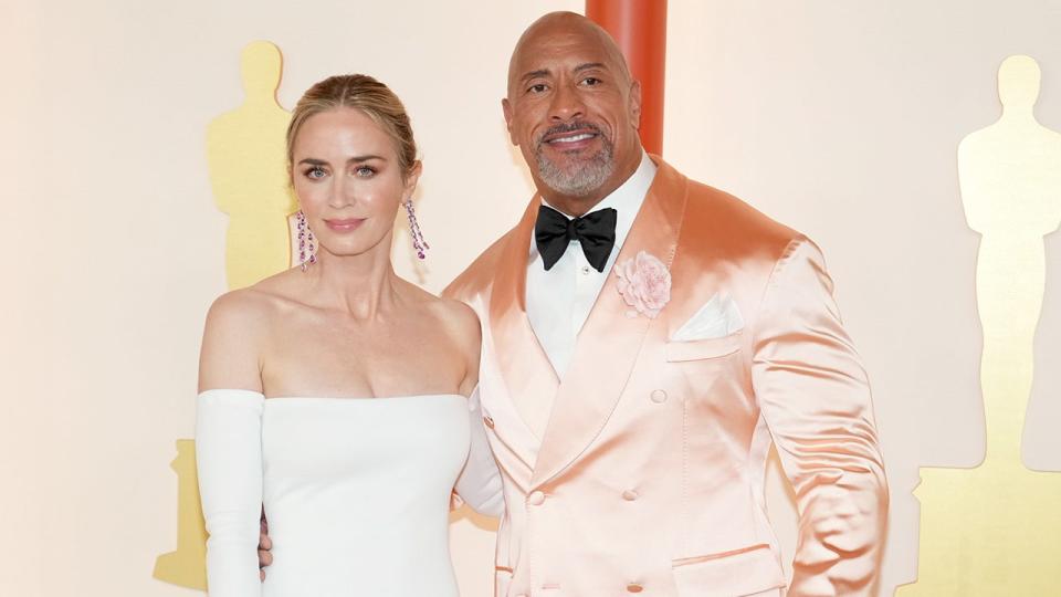 Emily Blunt in a white gown with long sleeves soft smiles on the Oscars carpet with Dwayne Johnson in a shiny peach suit