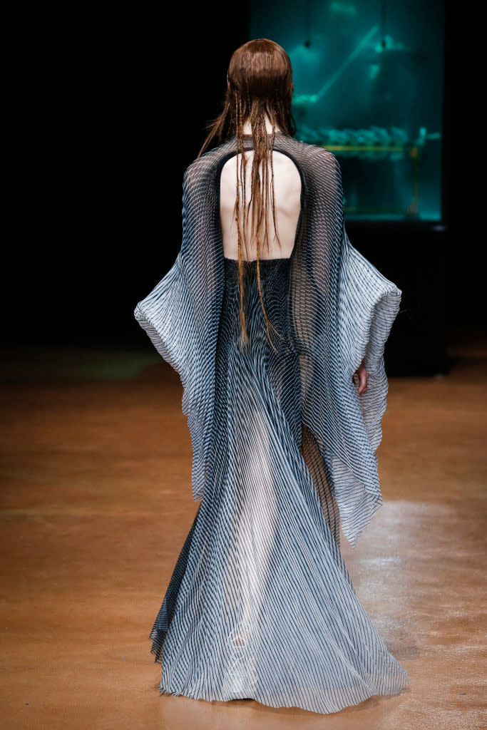 Model wearing a flowing, sheer dress with a striped pattern walks down a runway with their back facing the camera, hair appearing styled wet