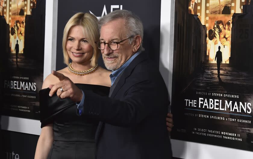 A blond woman in a black dress and a man with gray hair and glasses who is pointing in a black suit