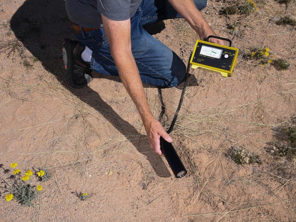 person crouching in jeans holds geiger counter little yellow box with wand detector over desert sand