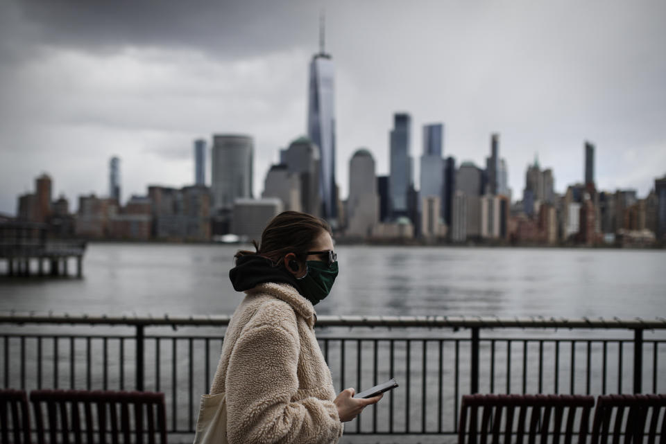 FILE- In this April 10, 2020 file photo a woman wearing a face mask sure to CVID-19 concerns walks along the Jersey City waterfront with the New York City skyline in the background. New York City may be the epicenter of the coronavirus outbreak but of the city's suburbs have been hit just just as hard. In some, there have been more fatalities per capita than in super-dense Manhattan. (AP Photo/John Minchillo File)