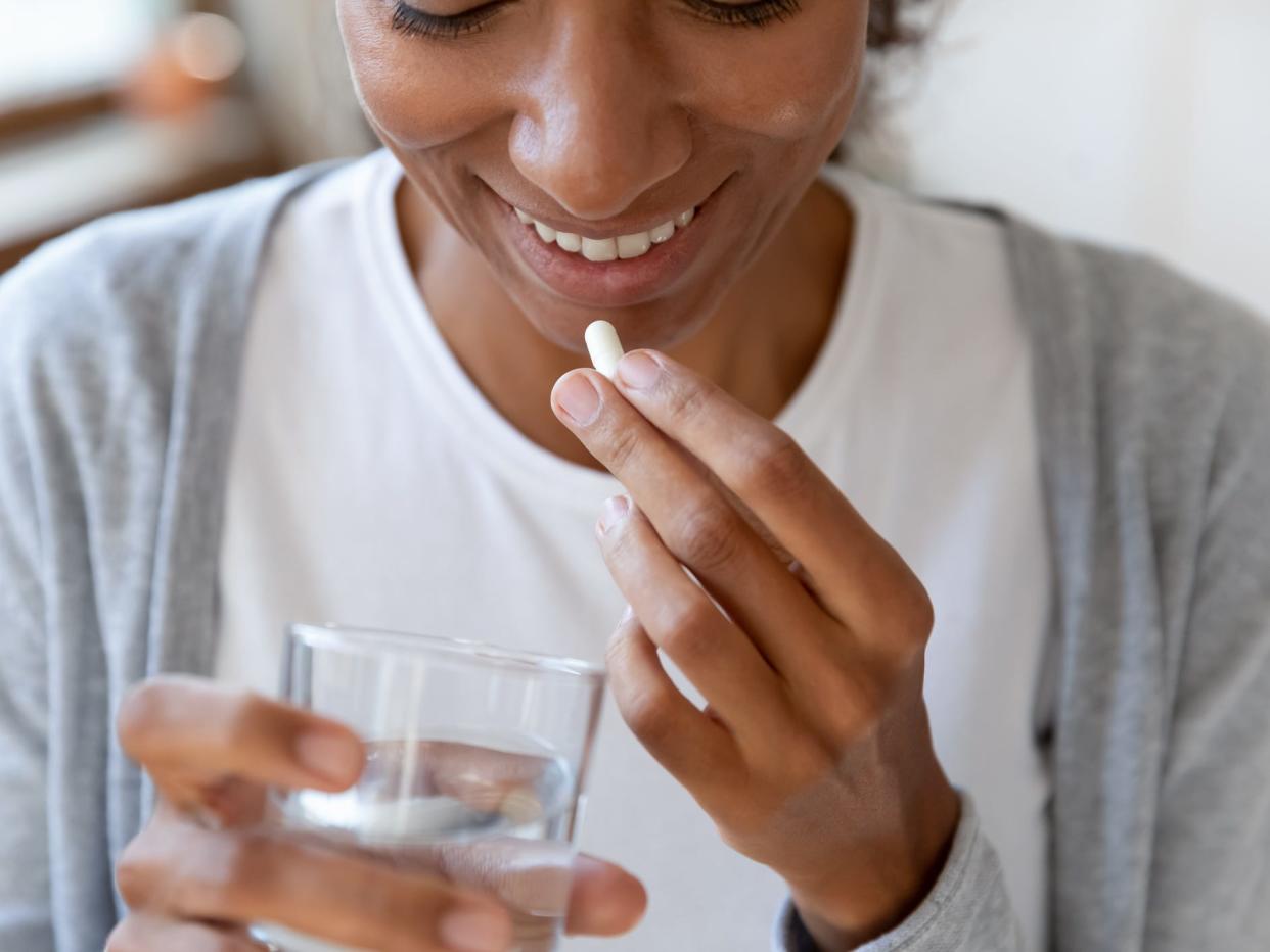 Image from the eyes down of a woman taking a pill and holding a glass of water. She has black eyelashes and smiles, looking down. She wears a grey sweater and white t-shirt. 