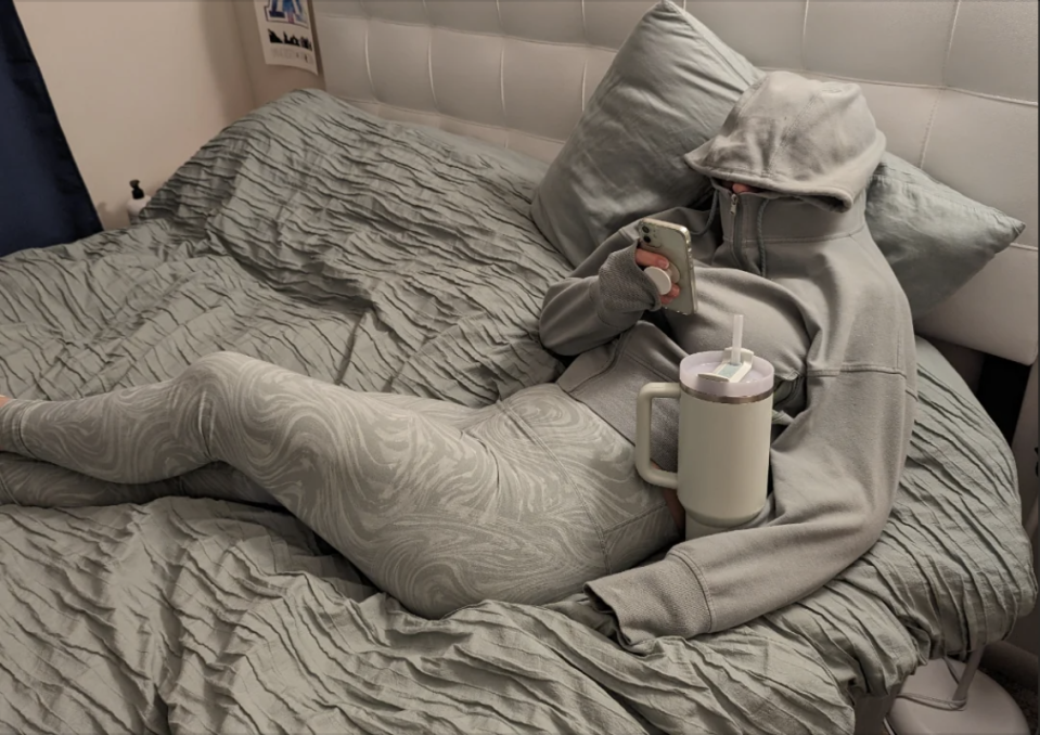 Person lying on a bed in a grey hooded tracksuit, holding a phone in one hand, with large cup and straw resting on their chest. Their face is covered by the hoodie