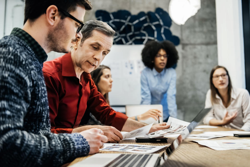 Two men are looking at documents in a business meeting. Women can be seen in the background talking to someone else.