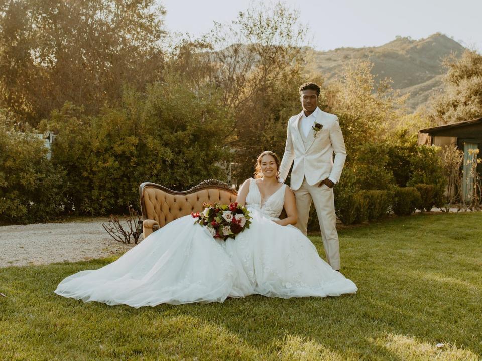 A bride sits on a couch outside and a groom sits next to her.