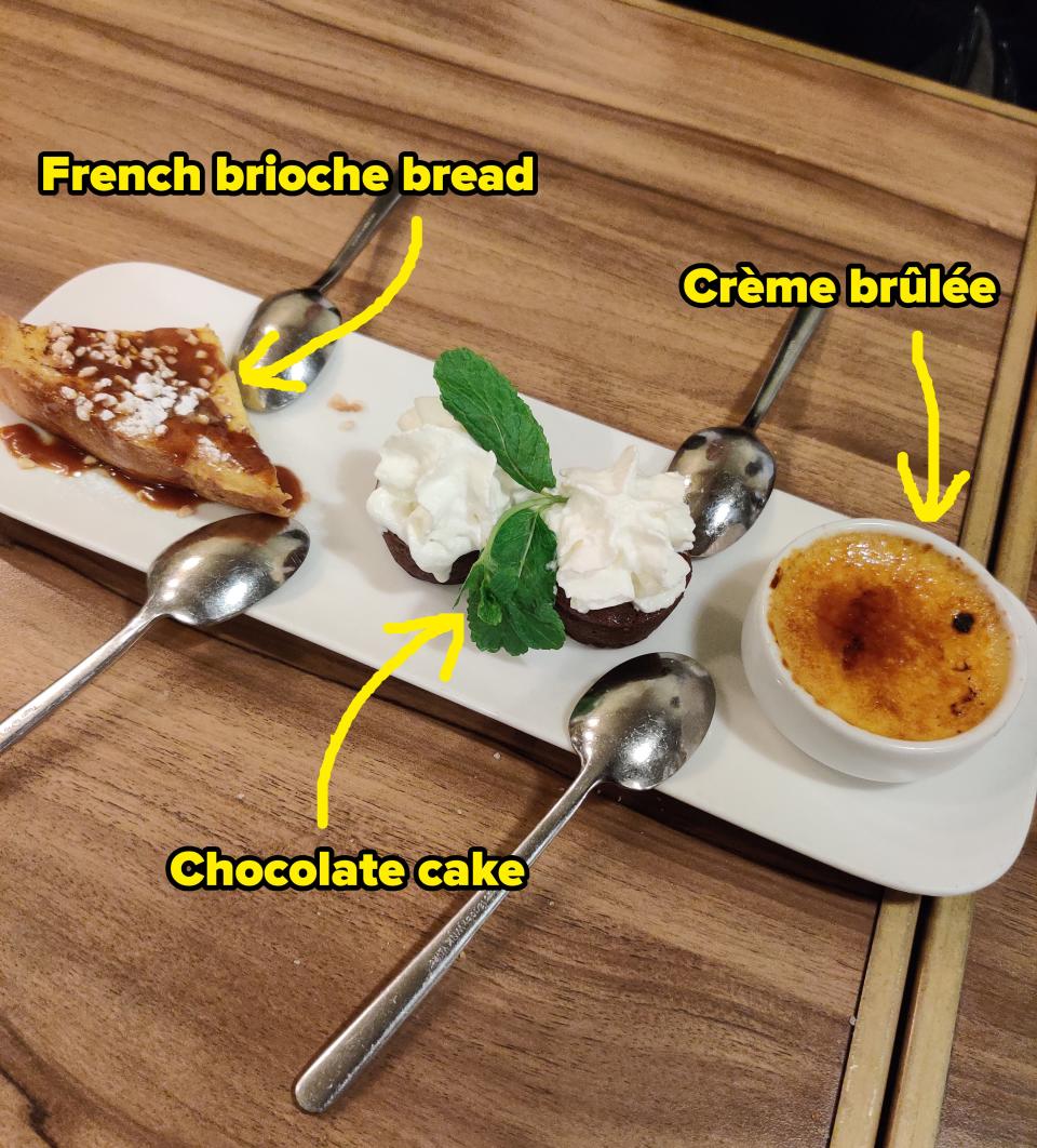 A tray with various desserts including a slice of cake, whipped cream, and a crème brûlée, with spoons, on a wooden table